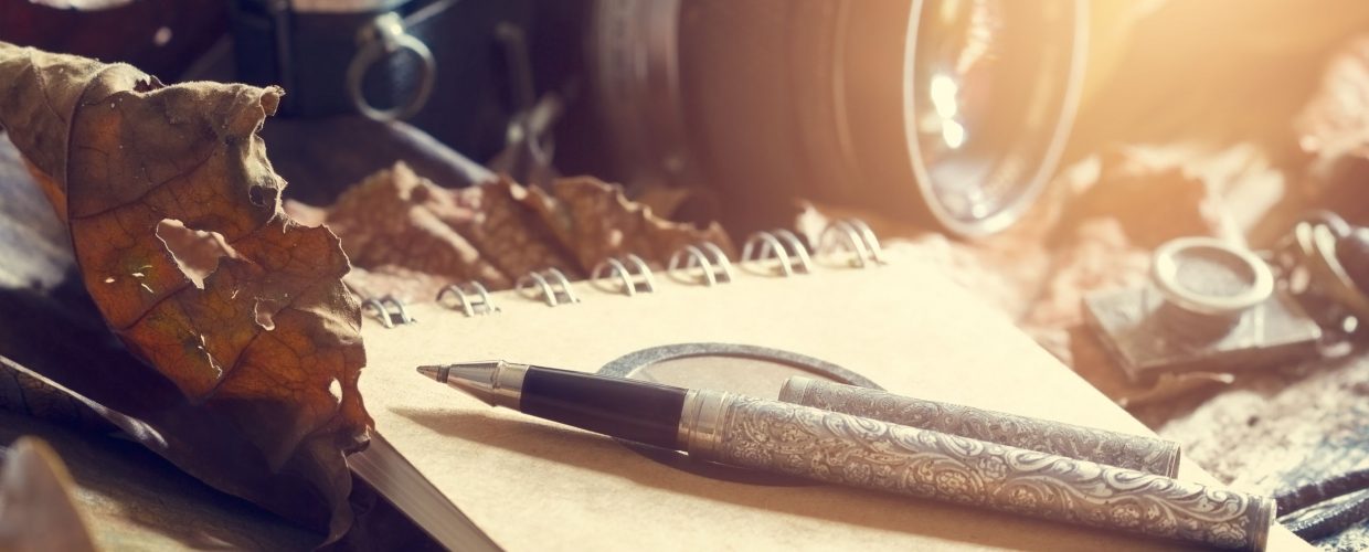 ABOUT US 48422629 - retro pen on old note pad and camera on dry leaf in jungle background, vintage color tone